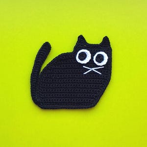 May include: A black crocheted cat-shaped item with large white eyes and white whiskers, set against a bright yellow-green background. The cat has a long tail and pointed ears. The item appears to be handmade.