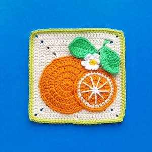 Puede incluir: Pieza cuadrada de ganchillo con un diseño de rodaja de naranja. Las rodajas de naranja son en tonos naranja con detalles blancos. También se incluyen hojas verdes y una pequeña flor blanca con centro amarillo. El cuadrado está bordeado con verde claro y crema.