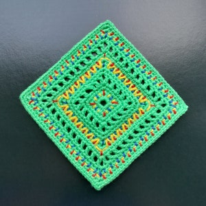 Könnte beinhalten: Ein quadratischer, grüner Häkelartikel mit einem geometrischen Lochmuster. Die Ränder und das Innere sind mit farbenfrohen Nähten in Rot, Gelb und Blau verziert. Der Artikel wird auf einer dunklen Oberfläche präsentiert.