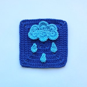 May include: A blue crocheted square featuring a blue crocheted cloud with raindrops hanging from it. The cloud is in the centre of the square.
