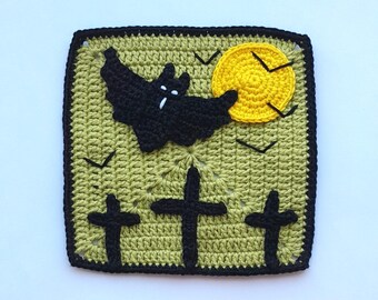 May include: A crocheted square featuring a Halloween theme. A black bat flies in front of a yellow moon, with three black crosses below. The background is a light green, and the border is black.