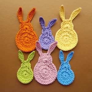 May include: Six crocheted bunny shapes in various colors, including orange, yellow, purple, pink, green, and blue. The bunnies are all made with a similar stitch pattern and have long ears.