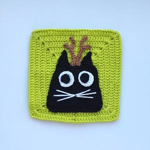 May include: A green crocheted square with a black cat face in the center. The cat has white eyes, a white mustache, and brown antlers.