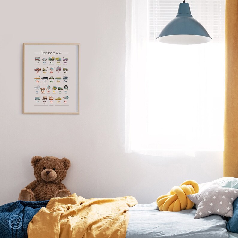 K&ouml;nnte beinhalten: Ein gerahmtes Poster mit dem Titel "Transport ABC" und Illustrationen verschiedener Fahrzeuge f&uuml;r jeden Buchstaben des Alphabets. Das Poster h&auml;ngt an einer wei&szlig;en Wand in einem Schlafzimmer mit einem Bett, einem Teddyb&auml;r und einem gelben Kissen.