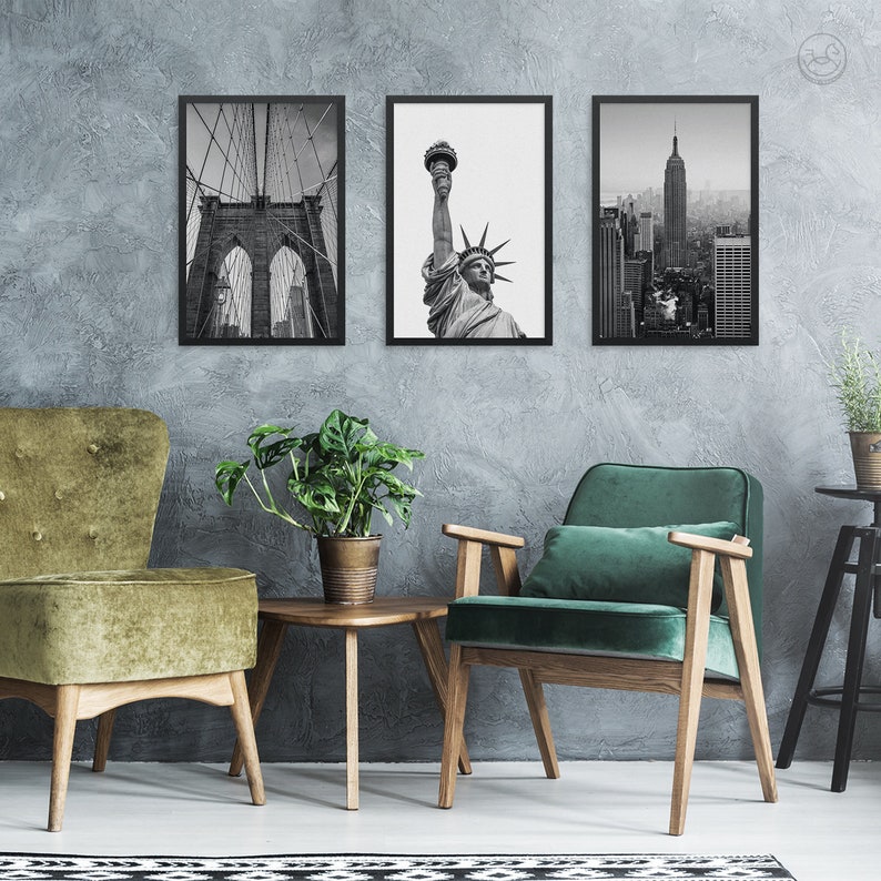 May include: Three black and white framed photographs of iconic New York City landmarks: the Brooklyn Bridge, the Statue of Liberty, and the Empire State Building. The photos are hanging on a gray wall above two green velvet armchairs and a wooden coffee table.