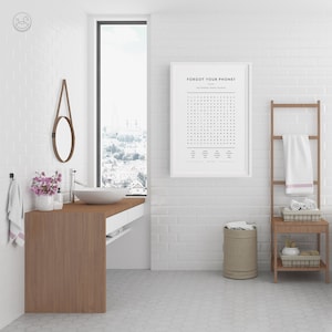 Op de afbeelding: Een witte badkamer met een witte bakstenen muur, een houten wastafel met een wit aanrecht, een ronde spiegel, een wit ingelijst print met de tekst "Forgot Your Phone!" en een woordzoeker, een bruine handdoekrek met een witte handdoek, een bruine kruk met een geweven mand en een beige wasmand.
