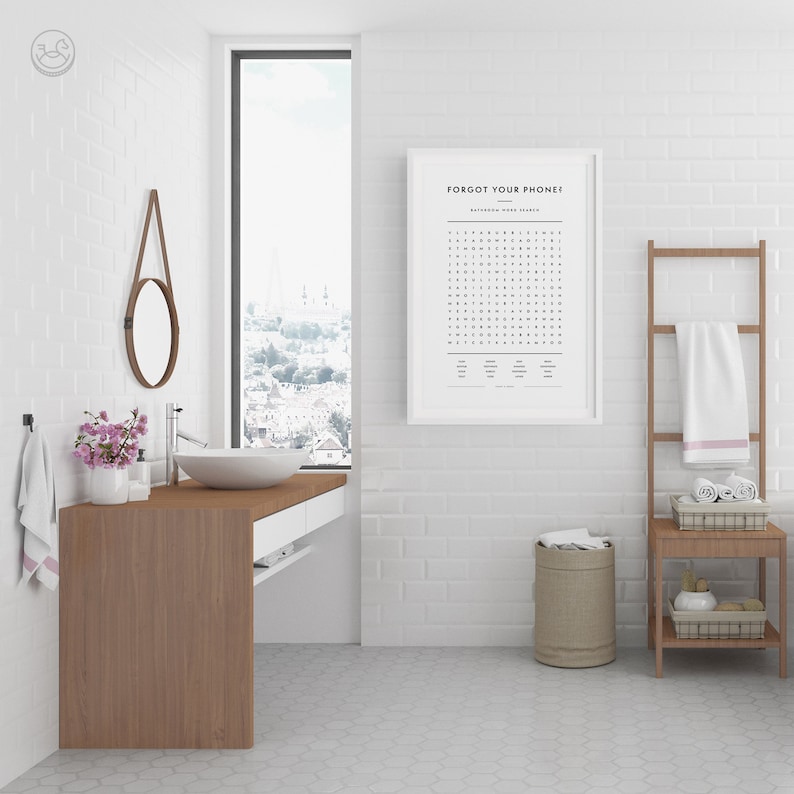 Op de afbeelding: Een witte badkamer met een witte bakstenen muur, een houten wastafel met een wit aanrecht, een ronde spiegel, een wit ingelijst print met de tekst "Forgot Your Phone!" en een woordzoeker, een bruine handdoekrek met een witte handdoek, een bruine kruk met een geweven mand en een beige wasmand.