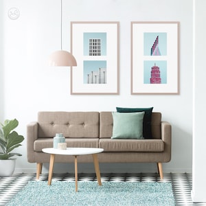 May include: A brown sofa with two green and teal throw pillows sits in front of a white wall with two framed prints of abstract cityscapes. A white coffee table with wooden legs sits in front of the sofa.