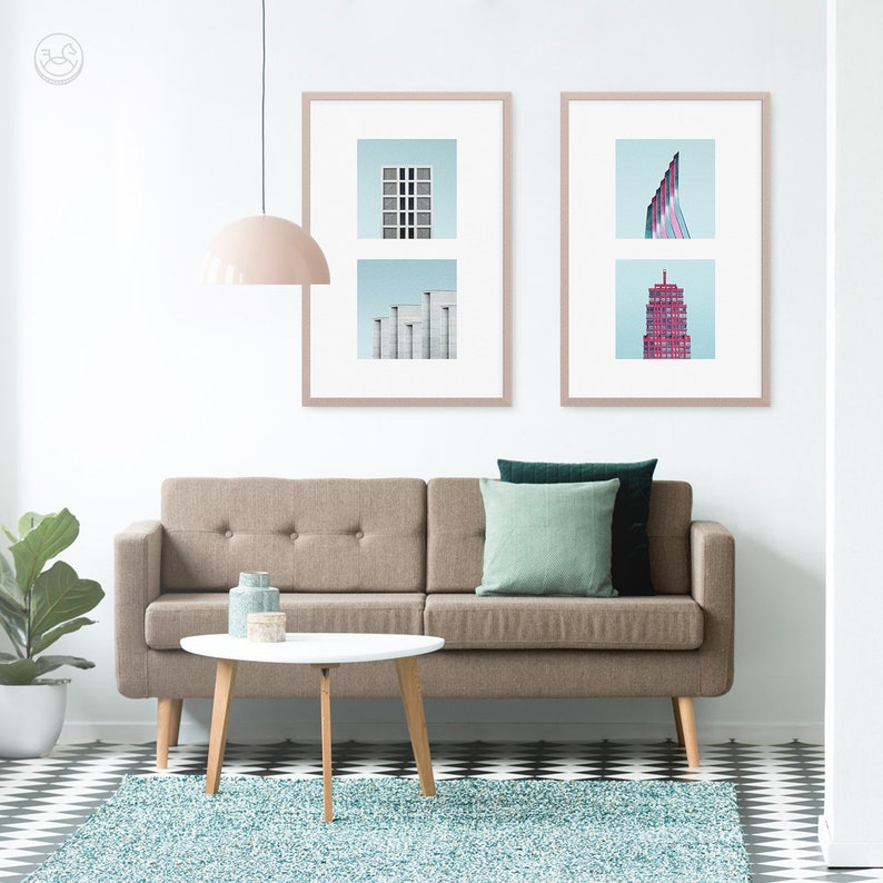 May include: A brown sofa with two green and teal throw pillows sits in front of a white wall with two framed prints of abstract cityscapes. A white coffee table with wooden legs sits in front of the sofa.