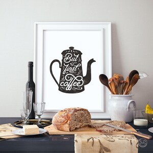 May include: A black and white illustration of a coffee pot with the text "But first, coffee" in a stylized font. The illustration is framed and hanging on a white wall.