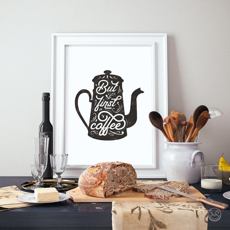 May include: A black and white illustration of a coffee pot with the text "But first, coffee" in a stylized font. The illustration is framed and hanging on a white wall.