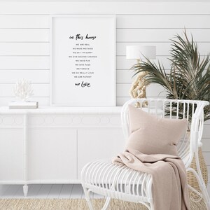 May include: A white framed print with black text that reads "in this house we are real, we make mistakes, we say I'm sorry, we give second chances, we have fun, we give hugs, we forgive, we do really loud, we are patient, we love". The print is hanging on a white shiplap wall. A white wicker chair with a pink throw blanket and pillow is in the foreground.