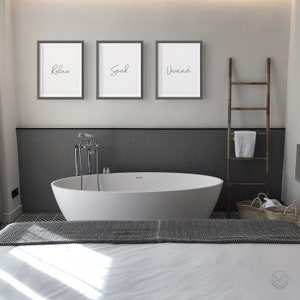 May include: Three framed prints with the words "Relax", "Soak", and "Unwind" hanging on a white wall above a white bathtub in a bathroom. A wooden ladder with a white towel hanging on it is to the right of the bathtub.