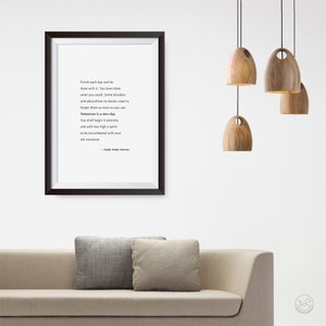 May include: A framed print with a black border displays a quote by Ralph Waldo Emerson. The quote is in black text on a white background. Wooden pendant lights hang from the ceiling. A beige sofa with two gray pillows is in the foreground.