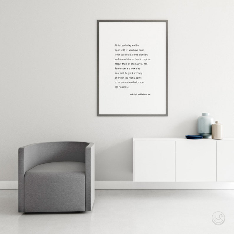 May include: A framed print with a quote by Ralph Waldo Emerson hangs above a white cabinet. The quote reads, "Finish each day and be done with it...Tomorrow is a new day." A gray armchair sits to the left. Decorative vases and a plate are on the cabinet.