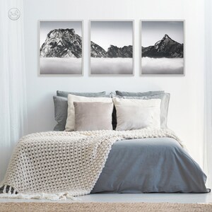 Pode incluir: Tr&ecirc;s fotografias em preto e branco emolduradas de montanhas com nuvens em primeiro plano. As fotografias est&atilde;o penduradas em uma parede branca acima de uma cama cinza com roupa de cama branca e um cobertor de croch&ecirc; branco.