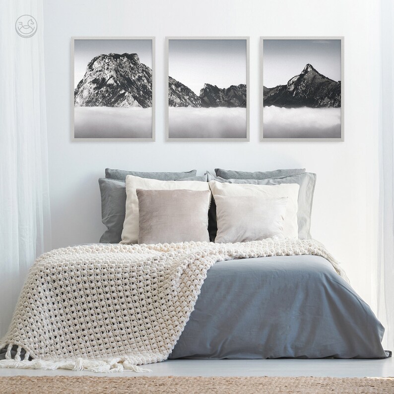 Pode incluir: Tr&ecirc;s fotografias em preto e branco emolduradas de montanhas com nuvens em primeiro plano. As fotografias est&atilde;o penduradas em uma parede branca acima de uma cama cinza com roupa de cama branca e um cobertor de croch&ecirc; branco.