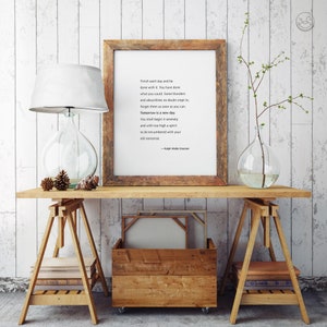 May include: A wooden framed print with a quote by Ralph Waldo Emerson sits on a wooden table. A glass lamp with a white shade and a clear glass vase with a plant are also on the table. A wooden crate and books are below.