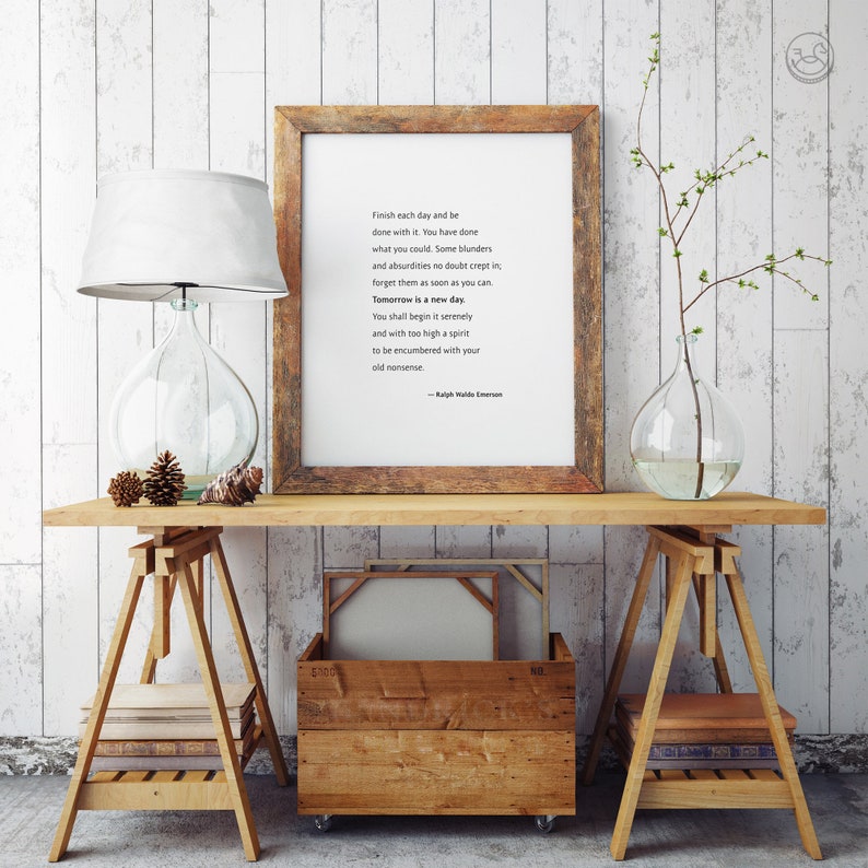 May include: A wooden framed print with a quote by Ralph Waldo Emerson sits on a wooden table. A glass lamp with a white shade and a clear glass vase with a plant are also on the table. A wooden crate and books are below.