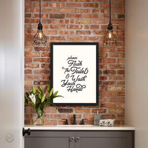 May include: A black framed bathroom sign with white text that reads "Please Flush the Toilet & Wash Your Hands". The sign is hanging on a brick wall with two black caged light fixtures above it.