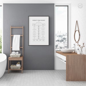 Op de afbeelding: Een ingelijst print met de tekst "Forgot your phone?" en een lijst met veelvoorkomende uitspraken en hun bijbehorende emoji. De print hangt aan een grijze muur in een badkamer met een witte tegelvloer, een houten wastafel en een wit bad.