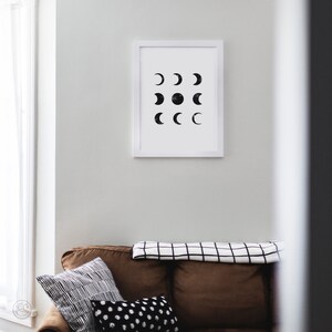May include: A white framed print with a black and white design of various phases of the moon. The print is hanging on a white wall above a brown couch with black and white patterned pillows.