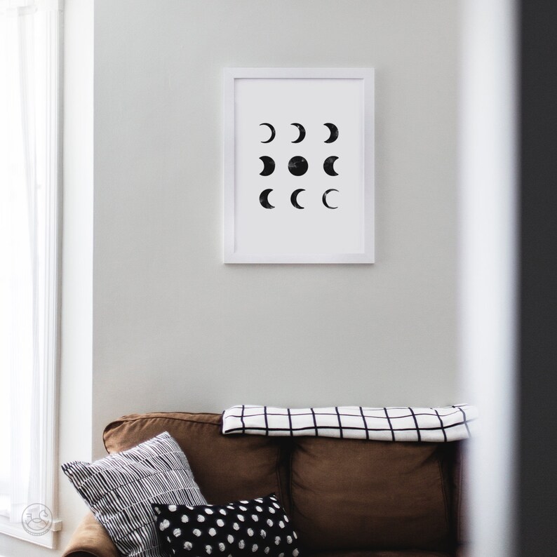 May include: A white framed print with a black and white design of various phases of the moon. The print is hanging on a white wall above a brown couch with black and white patterned pillows.