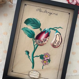 May include: Framed botanical print of an aubergine illustration. The artwork includes the word "Aubergine" in script, and the scientific name "Solanum melongena." The aubergine is depicted in purple, green, and pink, set against a beige background, within a black frame.