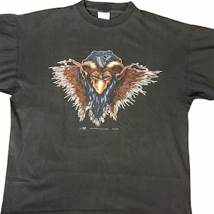 Puede incluir: Camiseta negra con un estampado gráfico de una criatura con cuernos, barba y alas. La criatura lleva un sombrero azul con un símbolo blanco.