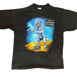 May include: A black short-sleeve t-shirt featuring a graphic of a lion holding a microphone. The text reads THE ROLLING STONES and BRIDGES TO BABYLON. The lion stands on a yellow desert landscape against a blue sky background on the front of the shirt.