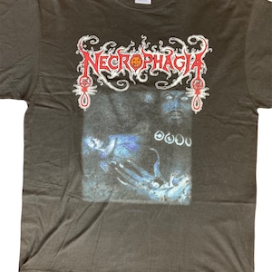 Puede incluir: Camiseta negra con un estampado gráfico de la banda "NECROPHAGIA" en letras rojas y blancas. El gráfico también incluye una imagen espeluznante de una cara y manos.