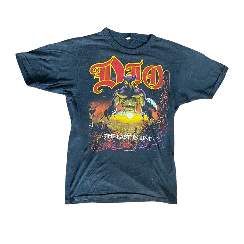 May include: Black t-shirt featuring a graphic print of a horned figure holding a staff. The text "DIO" is printed in red and yellow above the figure, and "THE LAST IN LINE" is printed below the figure.