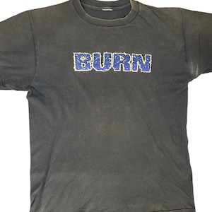May include: A dark gray t-shirt with the word "BURN" in a blue, textured font. The shirt has a crew neck and short sleeves. The fabric appears worn, giving it a vintage look.