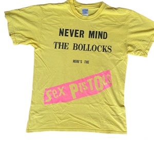 Könnte beinhalten: Gelbes T-Shirt mit schwarzem Text, der "Never Mind The Bollocks Here's The Sex Pistols" in einem rosa diagonalen Streifen lautet.