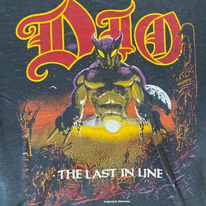 May include: A black t-shirt featuring a graphic print of a purple and gold demon with horns and chains. The text "DIO" is in red and yellow letters above the demon. The text "THE LAST IN LINE" is below the demon. The copyright text "&copy; 1984 N.E.T.T. PRODUCTIONS" is at the bottom of the shirt.