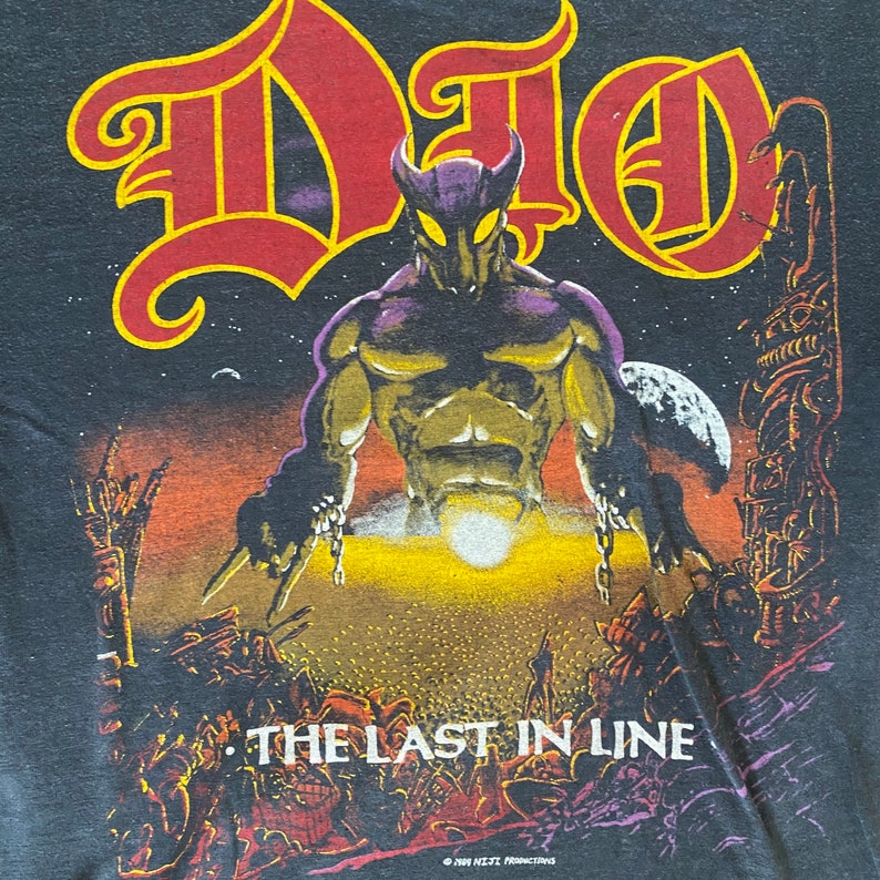 May include: A black t-shirt featuring a graphic print of a purple and gold demon with horns and chains. The text "DIO" is in red and yellow letters above the demon. The text "THE LAST IN LINE" is below the demon. The copyright text "&copy; 1984 N.E.T.T. PRODUCTIONS" is at the bottom of the shirt.