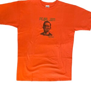 Op de afbeelding: Oranje T-shirt met een zwart-witte afbeelding van een man met een gasmasker. De tekst "PEARL JAM" en "BERLIN" staat op het shirt gedrukt.
