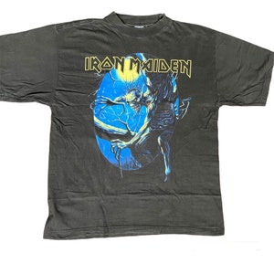 May include: A dark grey t-shirt with the band name "Iron Maiden" in yellow lettering. The graphic shows a skeletal creature with blue and yellow details against a blue backdrop.