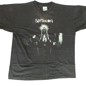 May include: Dark gray t-shirt featuring the band name "Satyricon" in white stylized text above a black and white graphic. The graphic depicts three figures with long hair and dark clothing, holding objects aloft.