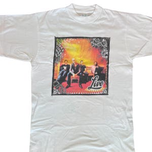 May include: White t-shirt featuring a graphic of a band sitting on a bench with a sunset background. The band's name, "Live", is printed in a cursive font. The shirt is a crew neck style.