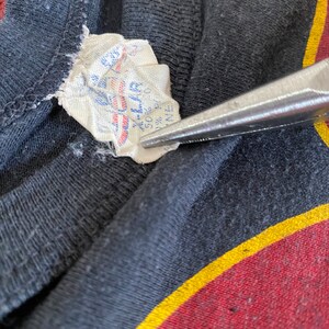 May include: A white tag with blue text sewn into a black garment. The tag reads "50% Cotton 50% Polyester Made in U.S.A."