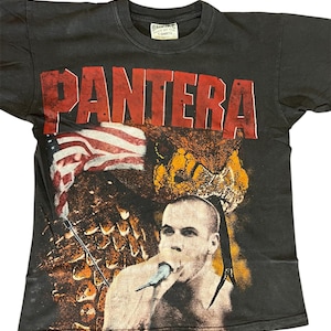 May include: A black short-sleeve t-shirt featuring the red text PANTERA at the top. The graphic includes a person singing into a microphone, a large snake head, and an American flag. The shirt has a white neck label with black text.