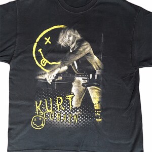 Op de afbeelding: Zwart T-shirt met een gele en zwarte afbeelding van Kurt Cobain die gitaar speelt. De afbeelding bevat de tekst "Kurt Cobain" en een smiley met een "X" over de ogen.