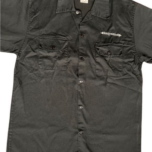 May include: Black short-sleeved button-up shirt with two chest pockets and the word "Electrolux" embroidered on the right chest pocket.