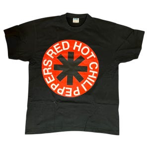 May include: Black t-shirt with a large red circle design. The words "PEPPERS RED HOT CHILI" are in white around the circle's edge. A black asterisk shape is in the centre. The shirt has a crew neck.