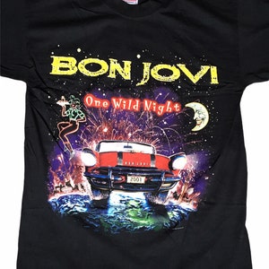 Puede incluir: Camiseta negra con un gráfico colorido de un coche volando por el aire con fuegos artificiales en el fondo. El texto "Bon Jovi One Wild Night" está impreso en amarillo en la camiseta.
