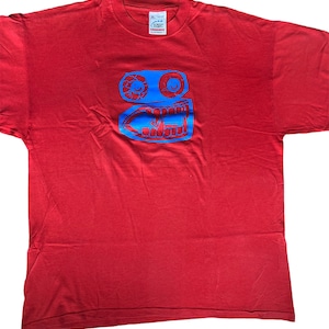 Può includere: Maglietta rossa con una grafica blu di un viso con due occhi e una bocca. La grafica è stilizzata e ha un aspetto da cartone animato. La maglietta ha un'etichetta Screen Stars.