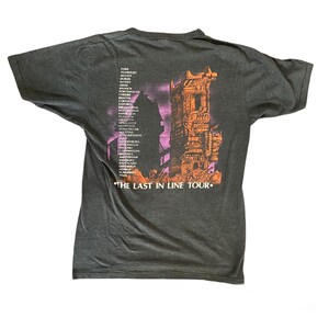 May include: A dark grey t-shirt featuring a graphic print of a futuristic city. The text "The Last in Line Tour" is printed at the bottom of the shirt. The city is depicted in shades of orange and pink.