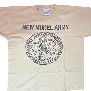 May include: Tan t-shirt with the text "NEW MODEL ARMY" above a circular, dark gray graphic. The graphic features an intricate design with swirling lines and a central floral element. The shirt has a crew neck.