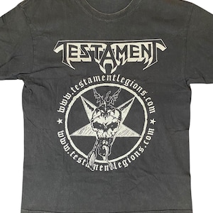 May include: Dark gray t-shirt featuring the band name "TESTAMENT" in white, stylized lettering. Below, a white pentagram design with a skull and the text "www.testamentlegions.com" encircles the star. A vintage band tee.